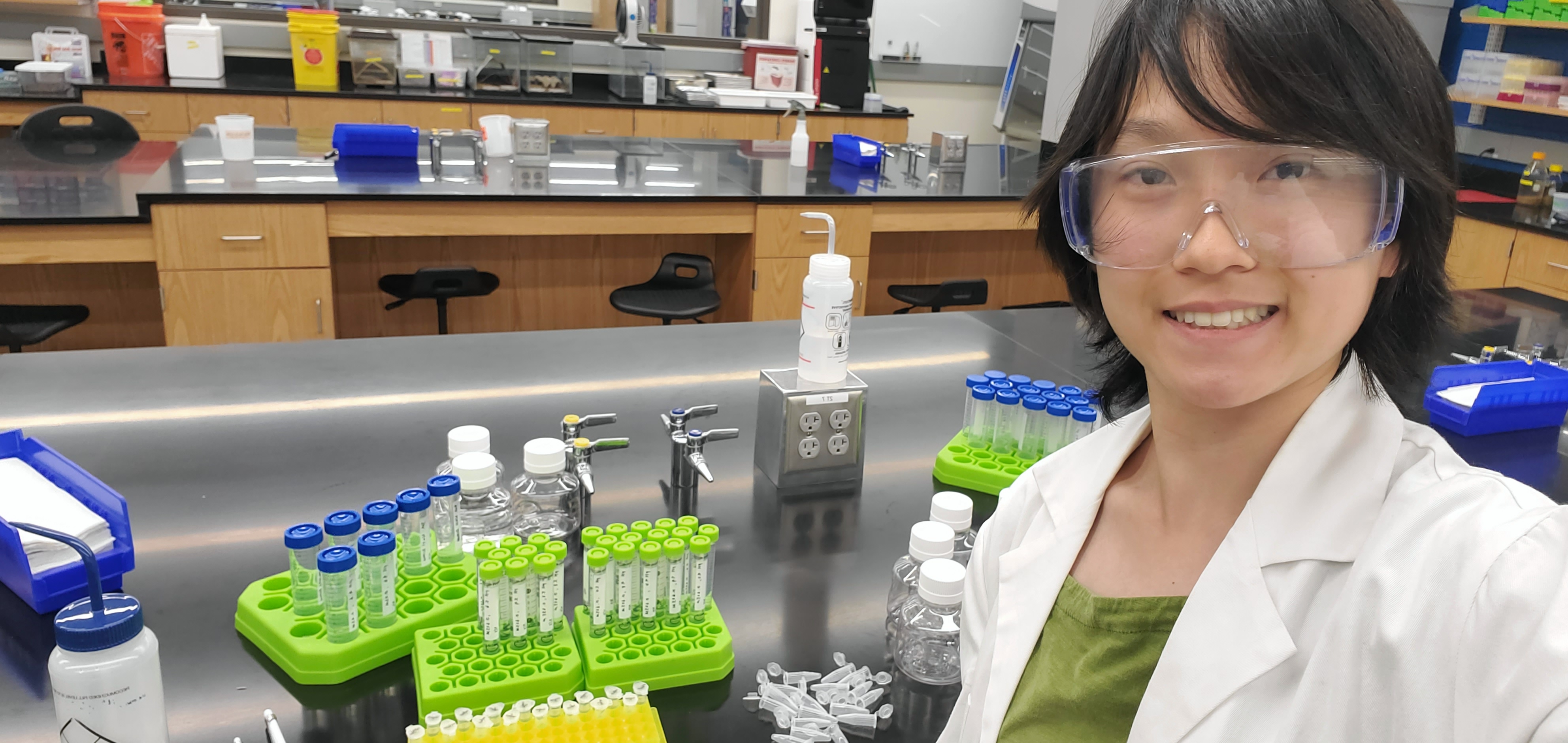 Susanna smiles at the camera, standing in front of lab equipment.