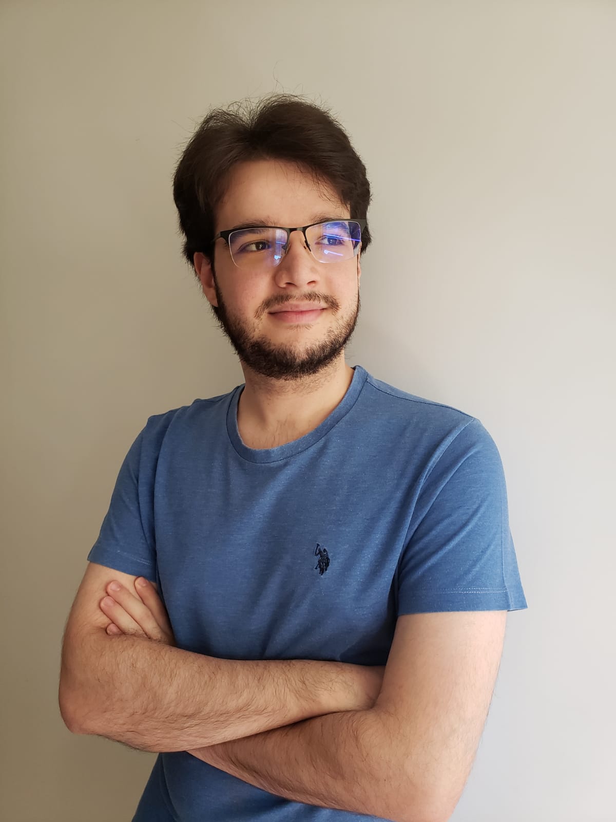 Carlos leans against a wall with arms crossed and a small smile on his face. He looks to his left and is wearing a blue shirt.